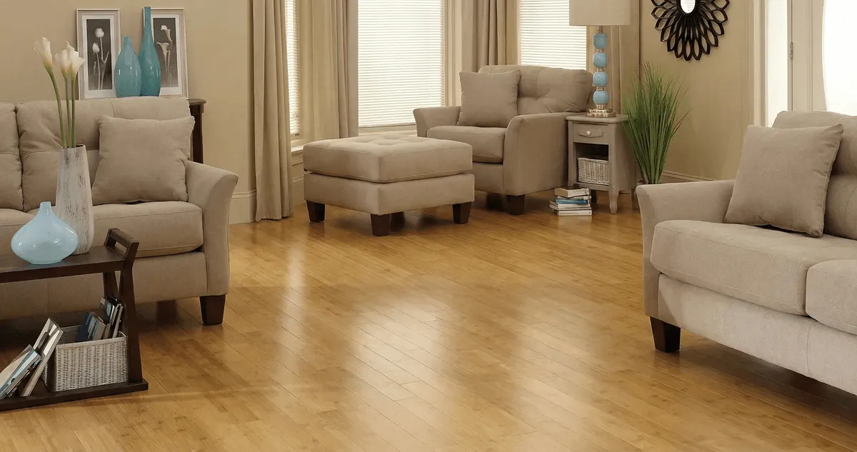 A living room with professionally installed bamboo flooring, beige sofas, a matching ottoman, a side table with a blue lamp, vases, framed pictures, and large windows with blinds and curtains in Dayton, OH